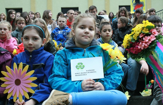 Grupa dzieci poubieranych w kurtki trzyma kwiaty, na głównym planie dziewczynka z kartką w dłoni