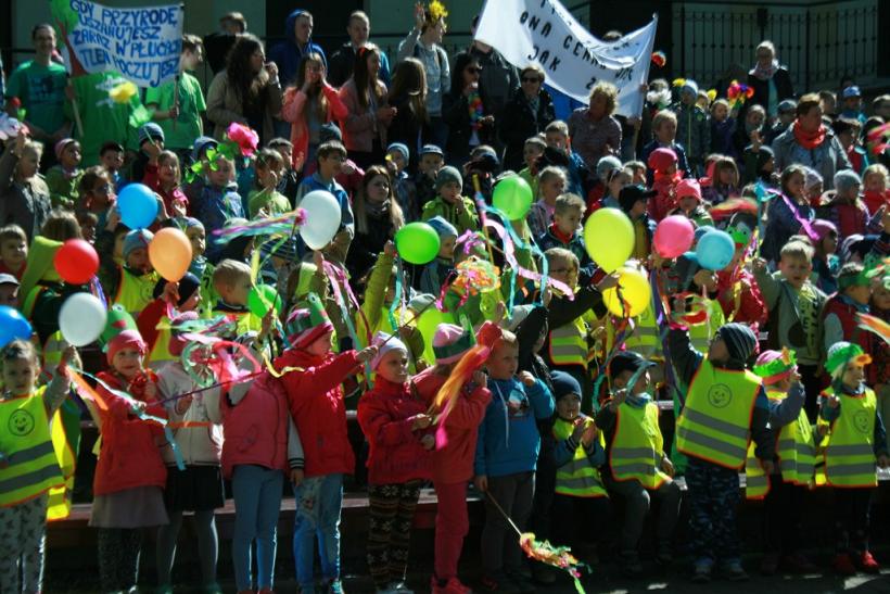 Dzieci w kurtkach i kamizelkach odblaskowych trzyma balony i różne inne dekoracje, w tyle stoją dorośli trzymający banery
