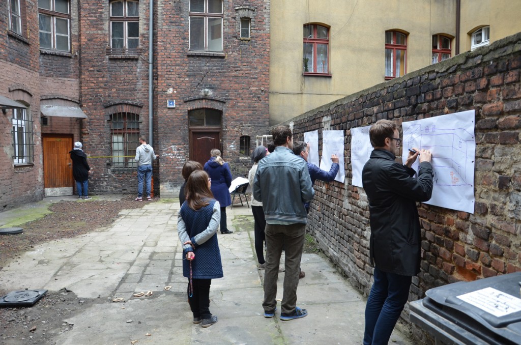 Ludzie rysujący po kartkach wywieszonych na ceglanym murze przy starych budynkach