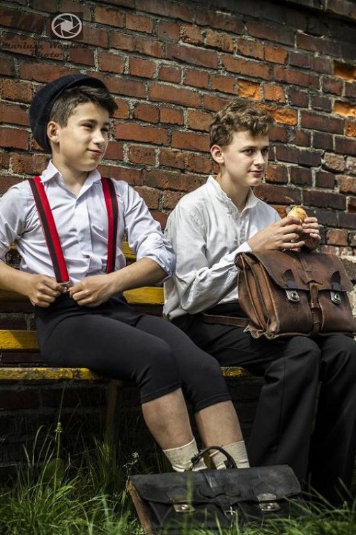 Dwoje elegancko ubranych chłopców z torbami siedzą na ławce przed ceglanym murem