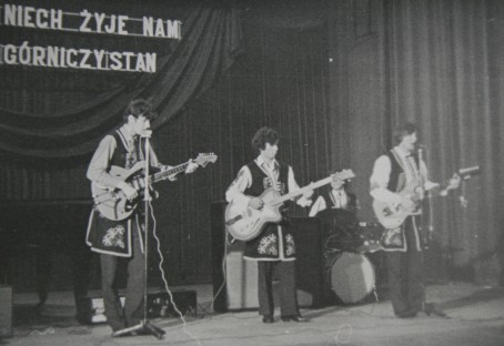 Troje ludzi ubranych w stroje ludowe grający na gitarach stoją na scenie.