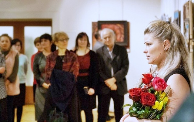 Po prawej stronie stoi kobieta z bukietem kwiatów, po lewej stronie widać grupę ludzi