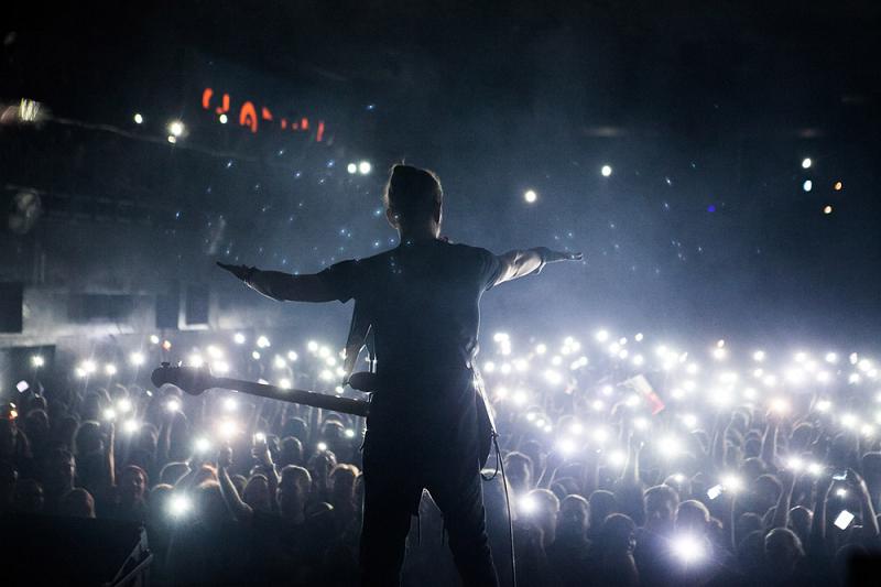 Osoba z rozłożonymi rękoma na scenie a za nią masa ludzi i ostrych piłek światła