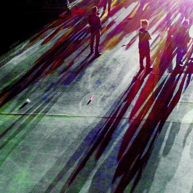 Tłum ludzi stojący przed słupem światła koncertu, za nimi długo rozciągające się cienie