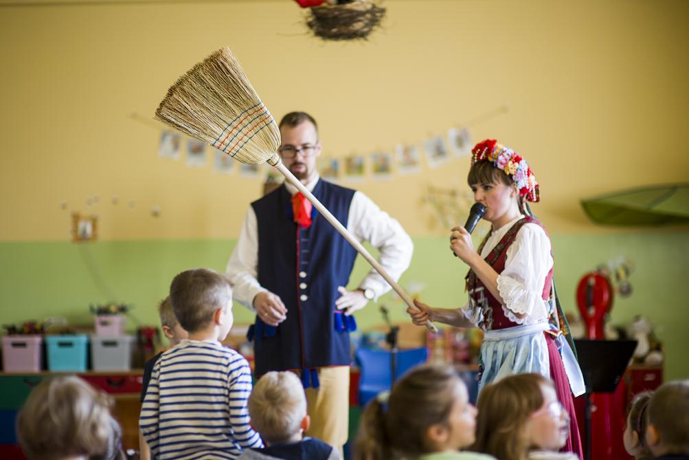 Tradycyjnie ubrany mężczyzna i kobieta z miotłą, widownia dzieci