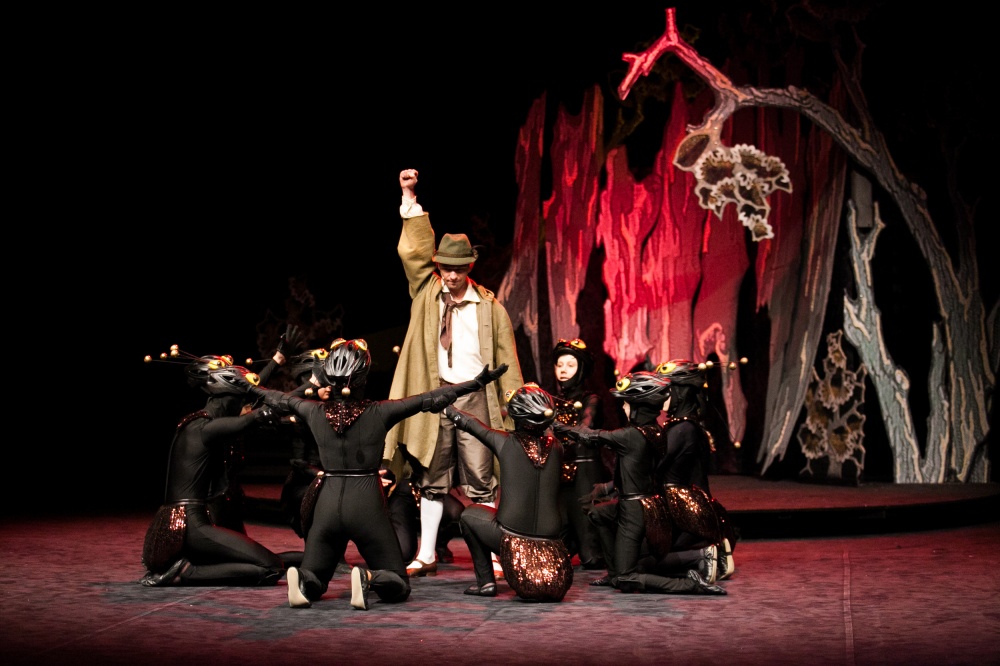 Mężczyzna ubrany na beżowo z skierowaną w górę pięścią stoi w kółku utworzony przez klękających aktorów ubranych w czerń