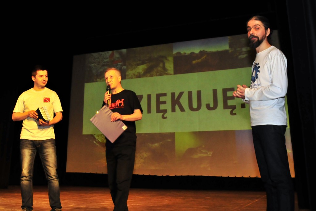 Trzech mężczyzn na scenie, jeden przemawia do mikrofonu i trzyma folder, za nimi na prezentacji napis 'Dziękuję.'