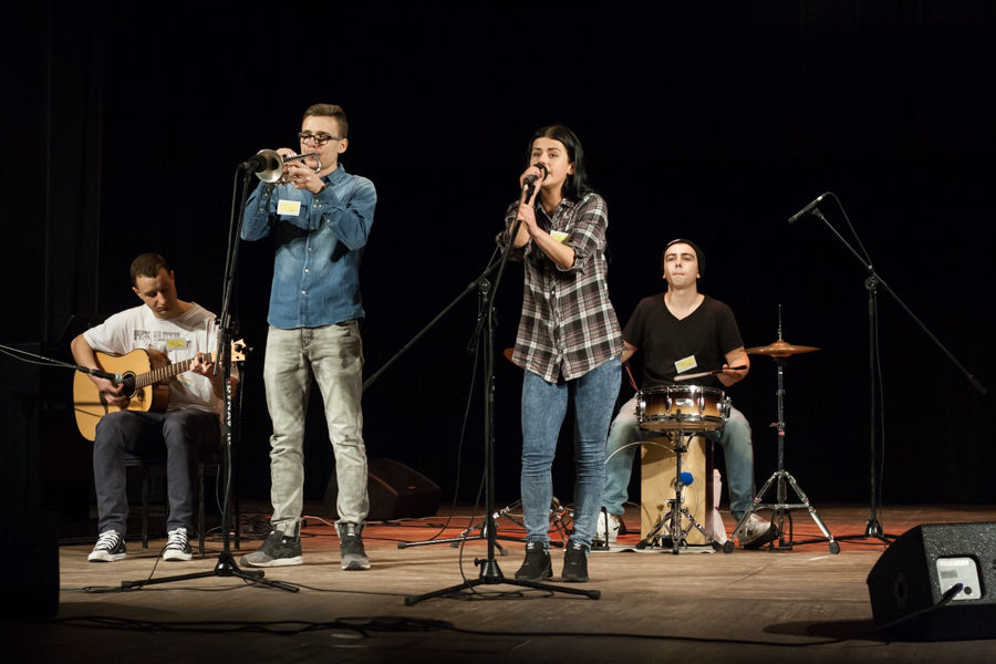 Para osób śpiewająca do mikrofonu, obok nich po dwóch stronach dwie osoby grające na gitarze i perkusji