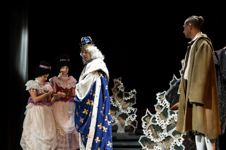 Aktor przebrany za króla w niebiesko-złotych szatach rozmawia z dwoma aktorkami i jednym aktorem