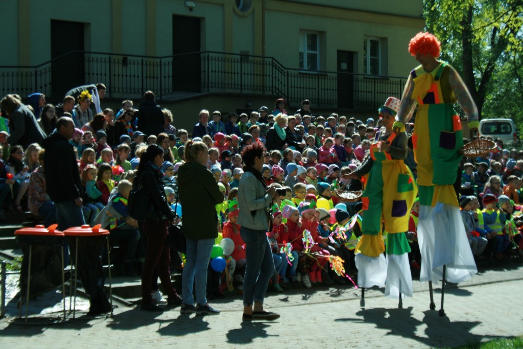 Widownia dzieci w parku trzymające patyki owinięte kolorową bibułą, wszystkie są ubrane w kamizelki odblaskowe, obok widowni dorośli opiekunowie, po chodniku idą dwie barwnie ubrane osoby na szczudłach