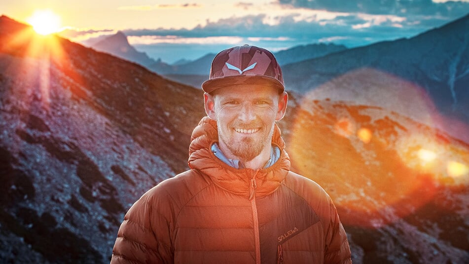 Uśmiechnięty mężczyzna w czapce na tle gór i zachodzącego słońca