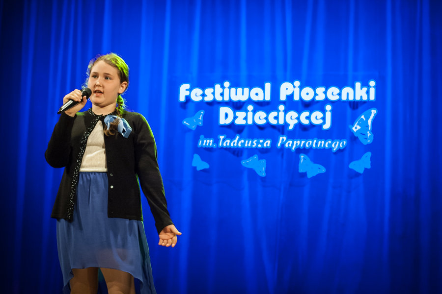 Dziewczyna w warkoczu śpiewająca do mikrofonu, za nią niebieska kurtyna z napisem 'Festiwal Piosenki Dziecięcej im. Tadeusza Paprotnego'