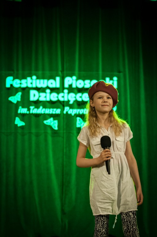 Dziewczynka w berecie trzymająca mikrofon, za nią zielona kurtyna z napisem 'Festiwal Piosenki Dziecięcej im. Tadeusza Paprotnego'