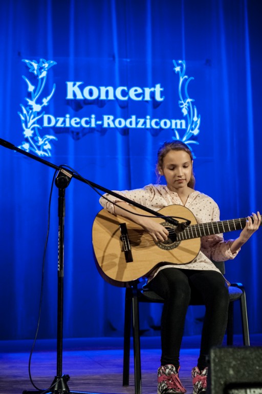 Dziewczynka grająca na gitarze, za nią niebieska kurtyna z napisem 'Koncert Dzieci-Rodzicom'