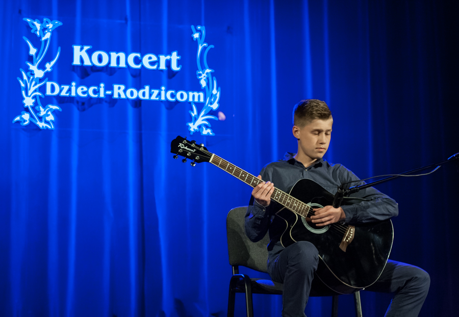 Chłopak grający na czarnej gitarze, za nim niebieska kurtyna z napisem 'Koncert Dzieci-Rodzicom'