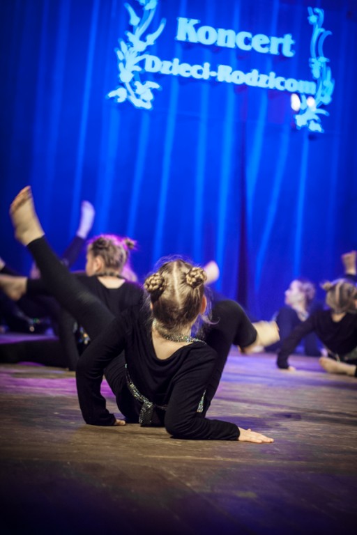 Dziewczynki ubrane w czarno-brokatowe stroje występują na scenie, za nimi niebieska kurtyna z napisem 'Koncert Dzieci-Rodzicom'