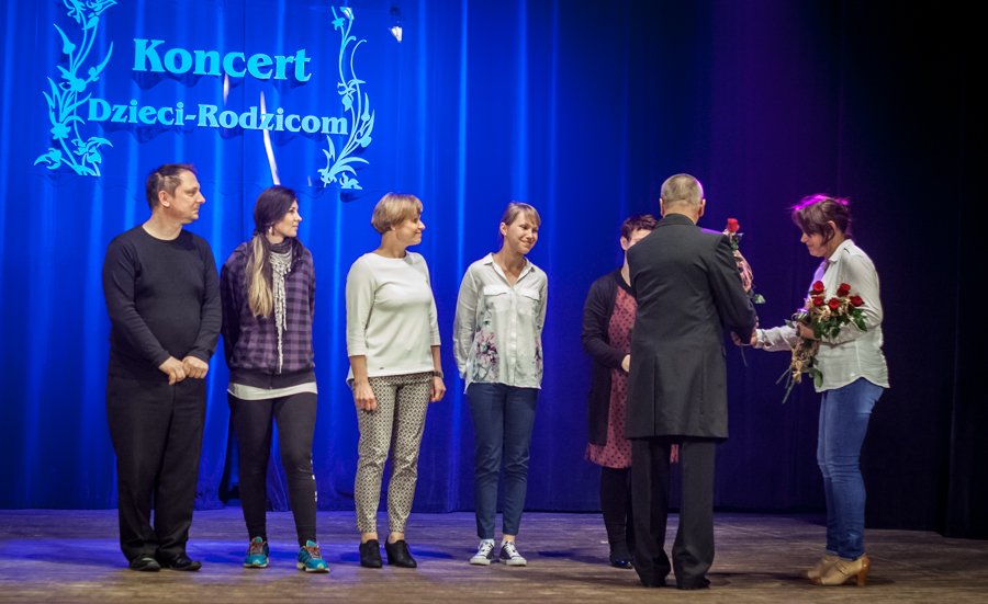 Elegancko ubrany mężczyzna wraz z jedną kobietą rozdaje nagrody osobom na scenie, za nimi niebieska kurtyna z napisem 'Koncert Dzieci-Rodzicom'