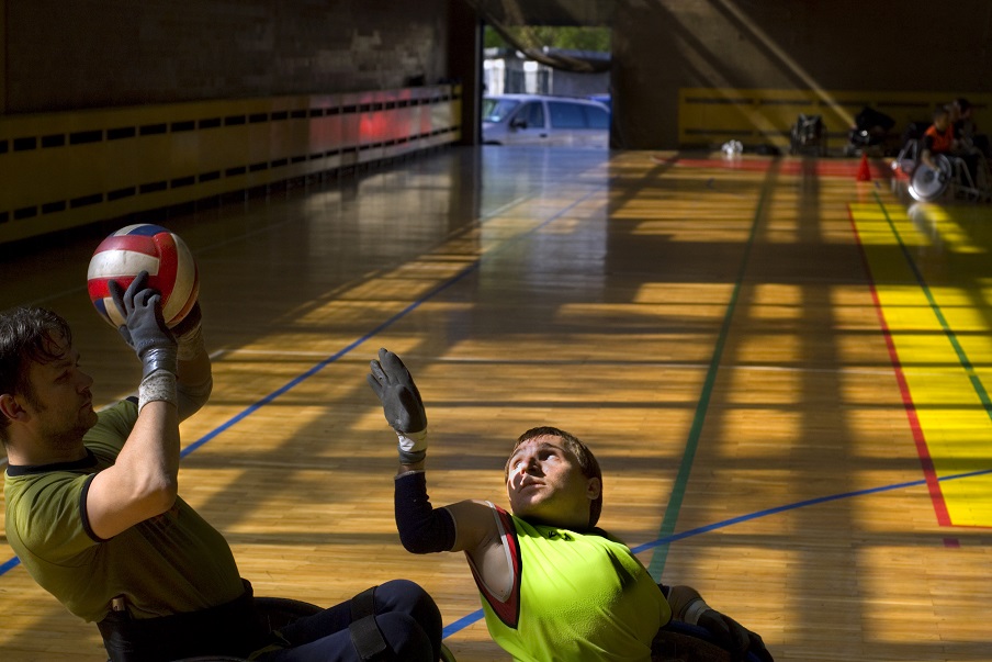 Mężczyzna trzyma piłkę do siatkówki, przed nim podnosząca rękę osoba niepełnosprawna na tle zakrytego boiska
