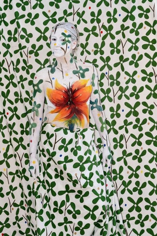 Osoba pomalowana w kwiatowy wzór na ścianie wtapiając się przy tym w tło, w centrum pomarańczowy kwiat wyróżniający się spośród zielonych, czterolistnych roślin