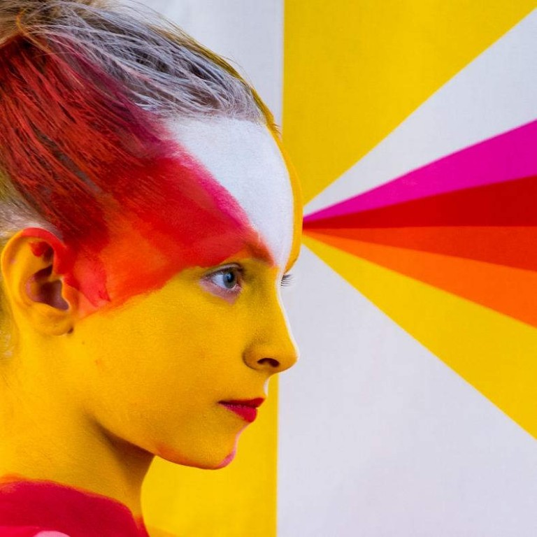 Dziewczynka pomalowana na biało-różowo-żółto, tło przedstawia te barwy (biały, różowy, czerwony, pomarańczowy i żółty) w formie geometrycznej