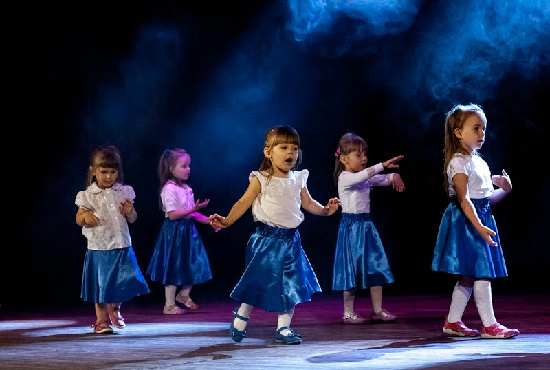 Małe dziewczynki w białych bluzkach i niebieskich spódniczkach występują na scenie