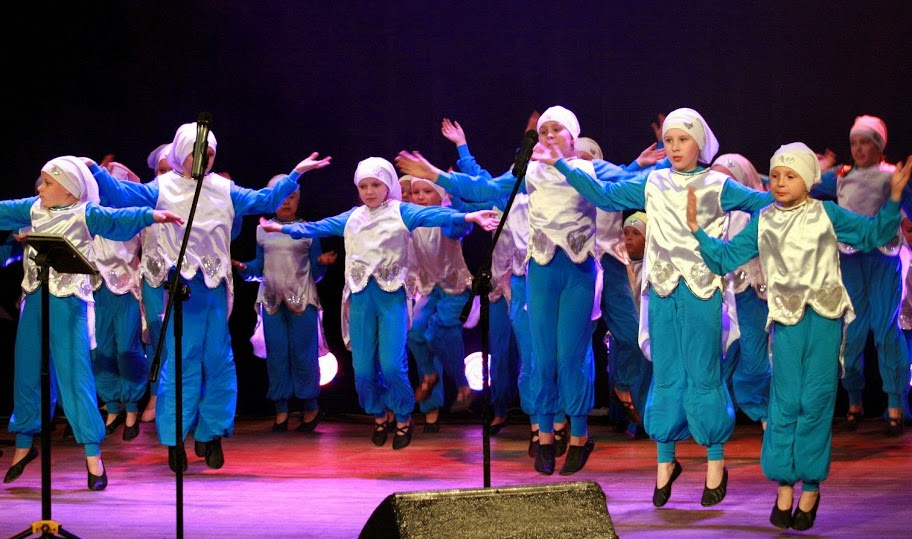 Dzieci ubrane w niebieskie spodnie, niebieskie bluzki, białe kamizelki i białe czapki występują na scenie