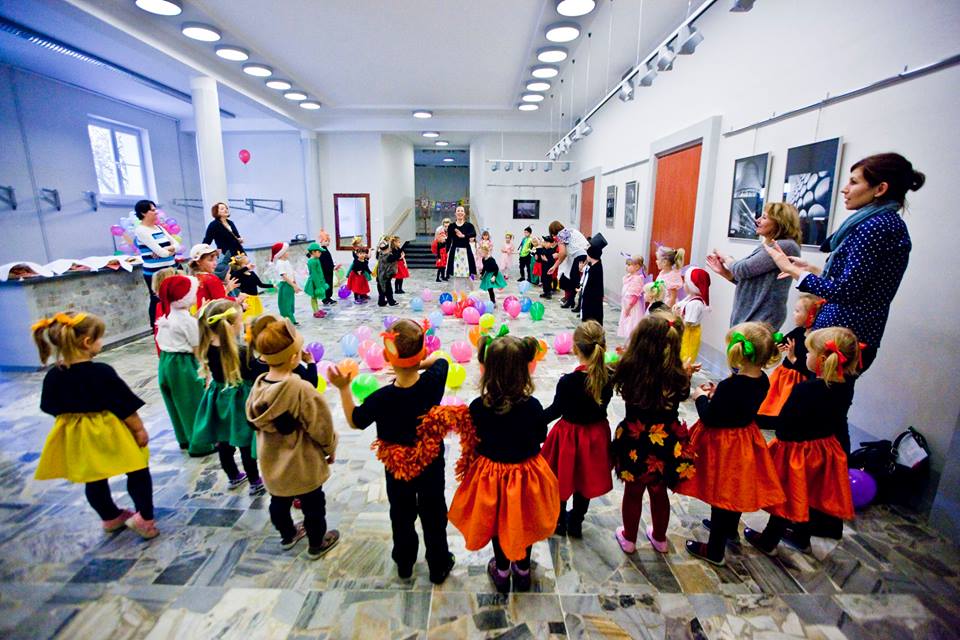Kolorowo ubrane dzieci stoją wraz z opiekunami w przestronnym pomieszczeniu