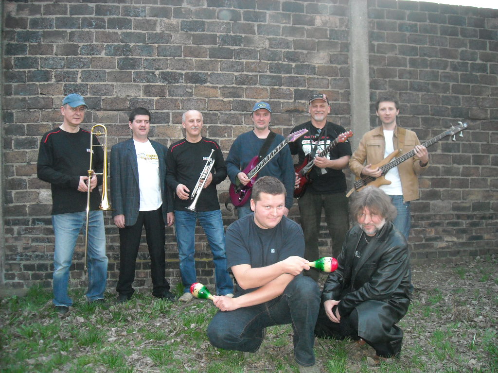 Zdjęcie grupowe ośmiu mężczyzn z instrumentami na tle ceglanego muru