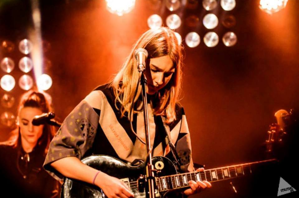 Dziewczyna grająca na gitarze, za nią pomarańczowe, sceniczne światło oraz inna osoba