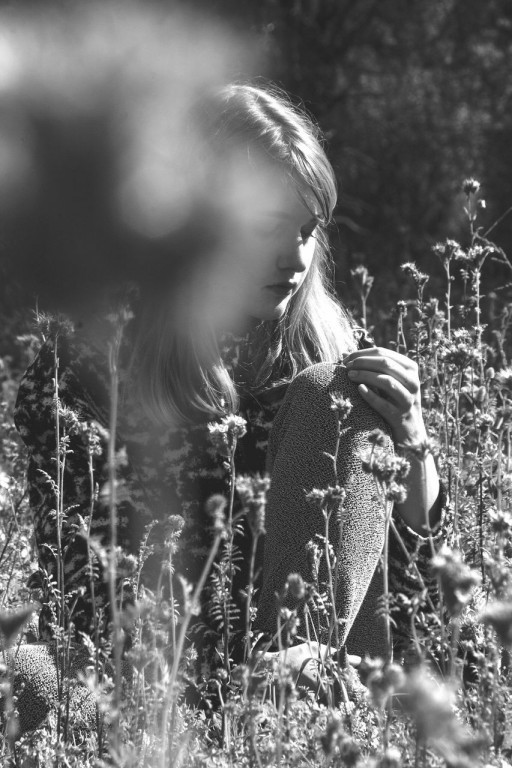 Czarno-białe zdjęcie kobiety siedzącej na polanie wśród wysokim kwiatów, jej sylwetkę zakrywa coś blisko kamery