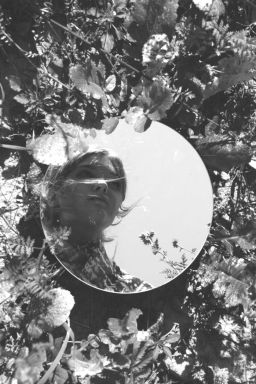 Czarno-białe zdjęcie odbicia kobiety w małym lusterku położonego na ziemi wśród roślin