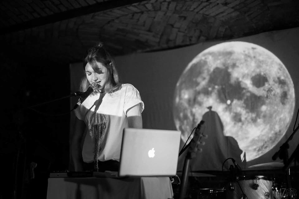 Czarno-białe zdjęcie kobiety przed mikrofonem, przed nią na biurku laptop z Apple, za nią prezentacja pokazująca księżyc w pełni