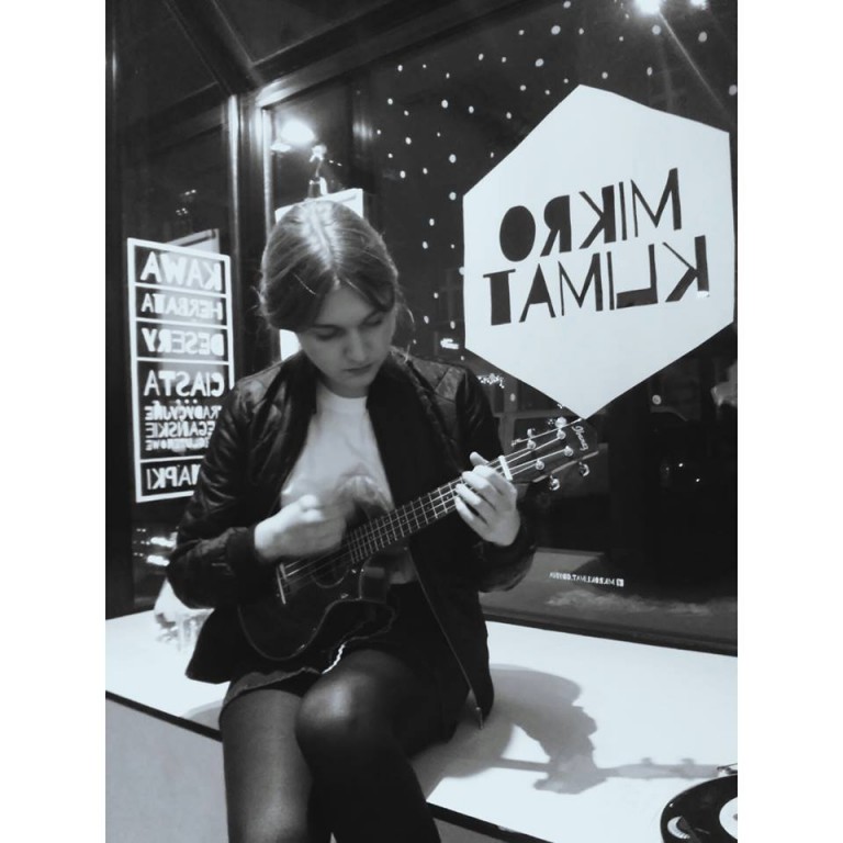 Czarno-białe zdjęcie kobiety grająca na gitarze przed naklejką na ścianie z napisem 'MIKRO KLIMAT'