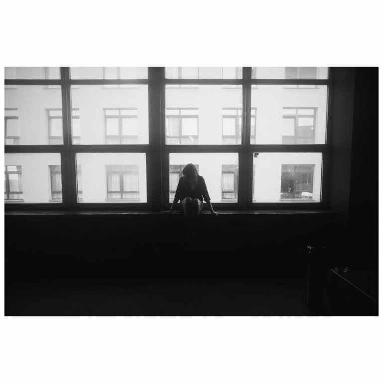 Czarno-białe zdjęcie kobiety siedzącej na parapecie przed dużym oknem