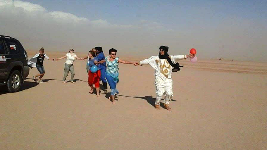 Grupa ludzi na pustyni trzyma się za ręce, idą ze strony czarnego samochodu po lewej