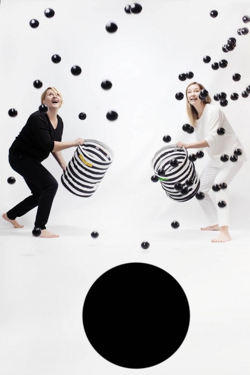 Dwie kobiety, jedna ubrana w całości na czarno, druga na biało, rozrzucają czarne kulki z koszyków w czarno-białe paski, pod nimi duże czarne koło