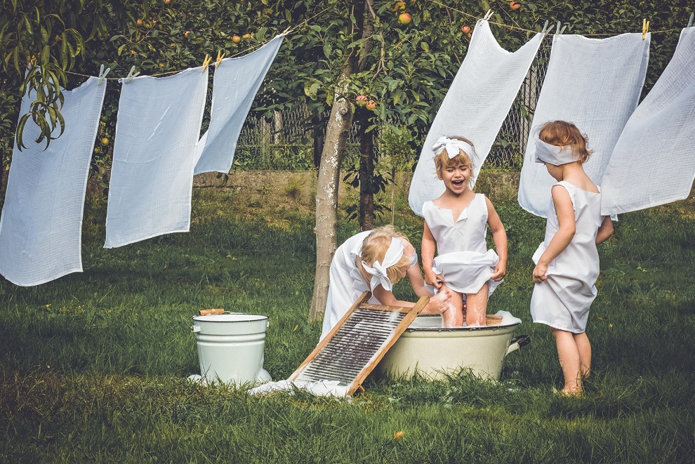 Trzy małe dziewczynki obok suszącego się prania, jedna w misce do prania, druga myje jej nogi