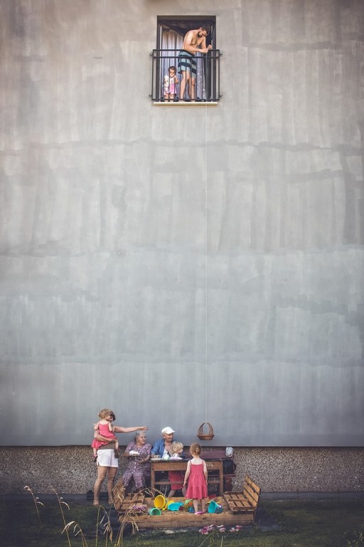 Mężczyzna z balkonu spuszczający na linie koszyk do dziadka, babci i matki z dziećmi siedzącymi przy stoliku