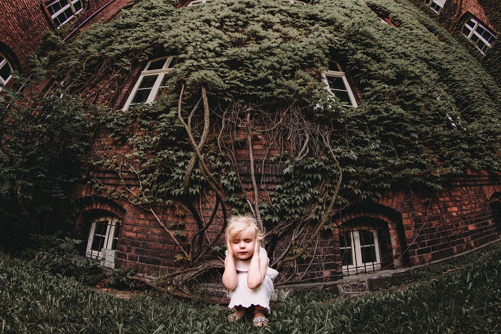 Klęcząca i zasłaniająca uszy dziewczynka na tle zarośniętego roślinnością budynku