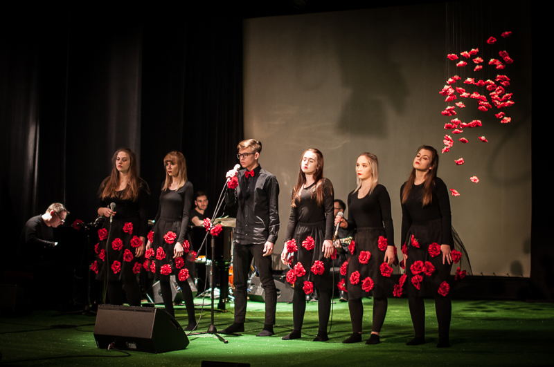 Przedstawienie, chłopak mówiący do mikrofonu i pięć kobiet w sukniach pokrytych różami obok