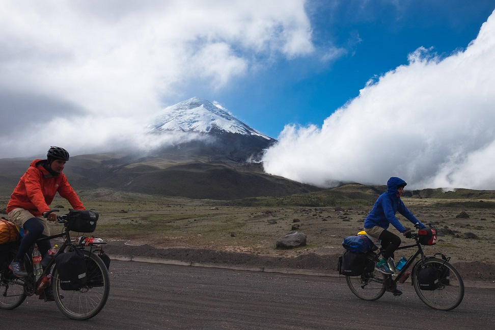 Dwoje ludzi na rowerach w kurtkach, z plecakami i bagażem. W tle góry i niskie chmury
