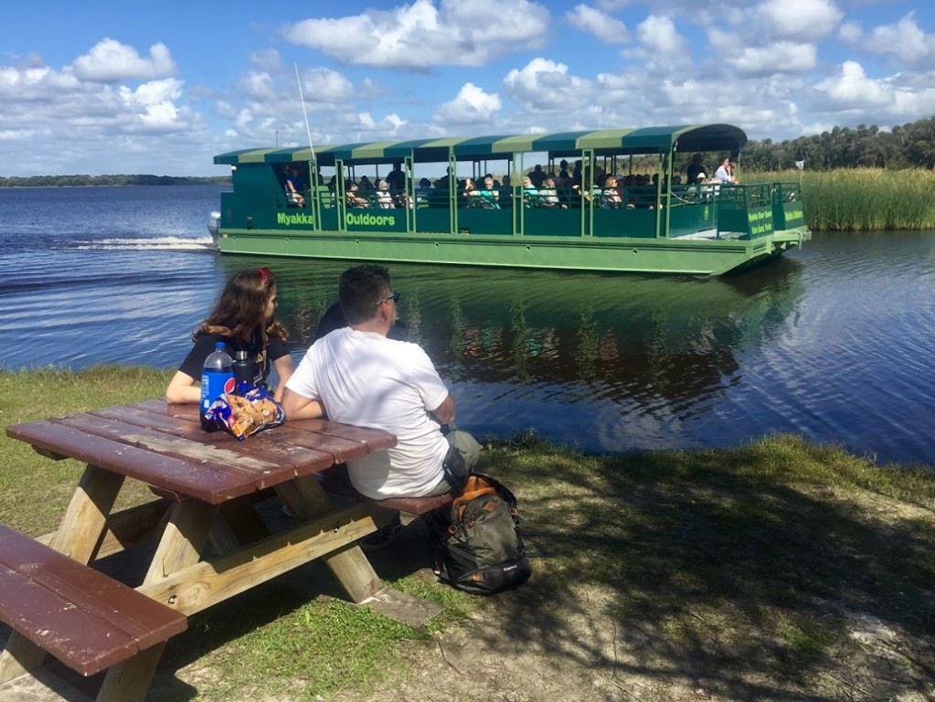 Mężczyzna i kobieta przy stole koło rzeki przez którą przepływa duża barka z turystami