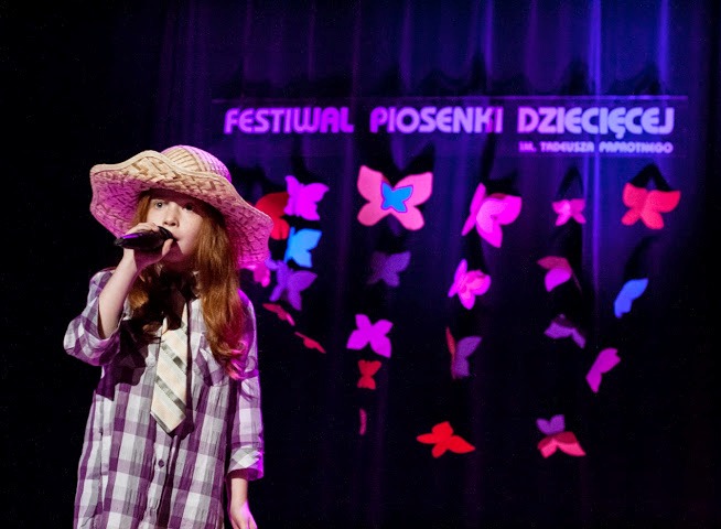 Dziewczynka w kapeluszu śpiewa na scenie, za nią napis 'Festiwal Piosenki Dziecięcej' i poprzypinane papierowe motyle