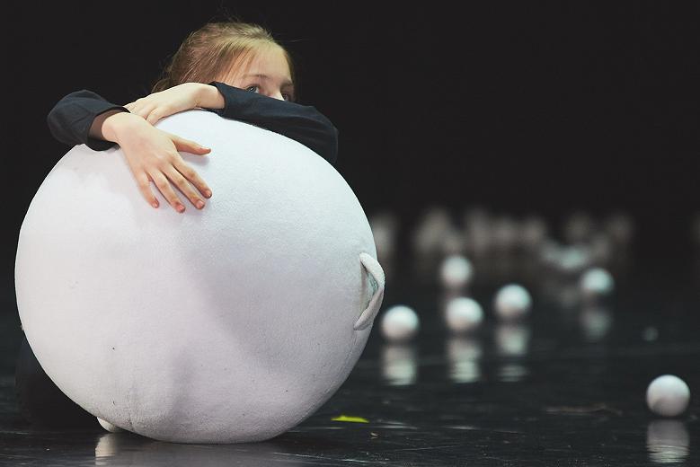Dziecko opierające się o dużą, białą kulę na scenie, za nim porozrzucane małe białe kulki