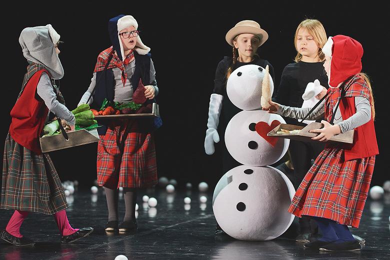 Dzieci na scenie poubierane w zimowe stroje, trzy niosą tacy z jedzeniem, dwie stoją przy sztucznym bałwanie