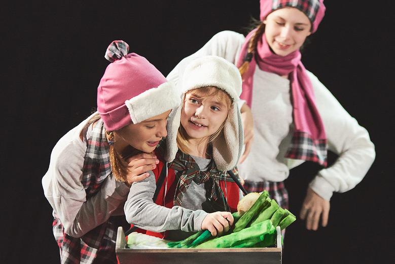 Troje dziecięcych aktorów zimowo poubieranych gra na scenie, jeden z nich trzyma tace z selerem