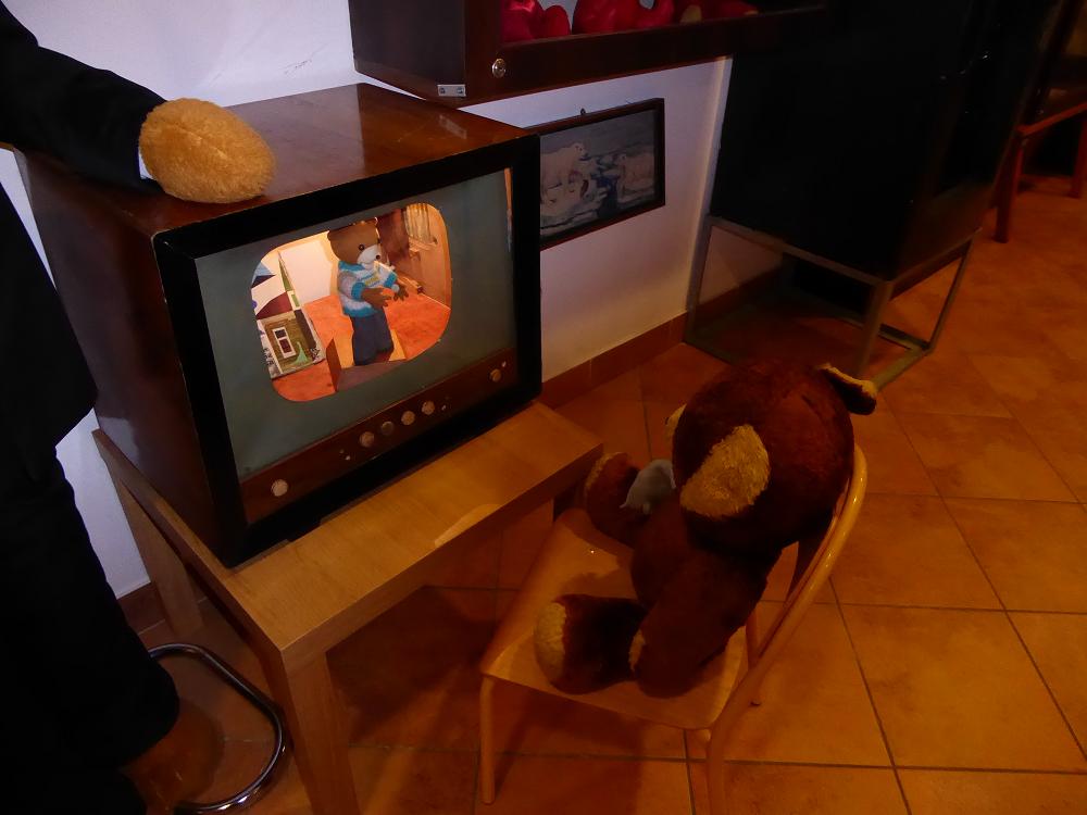 Stary telewizor, w nim puszczony film animowany, na krześle przed nim siedzi pluszowy miś