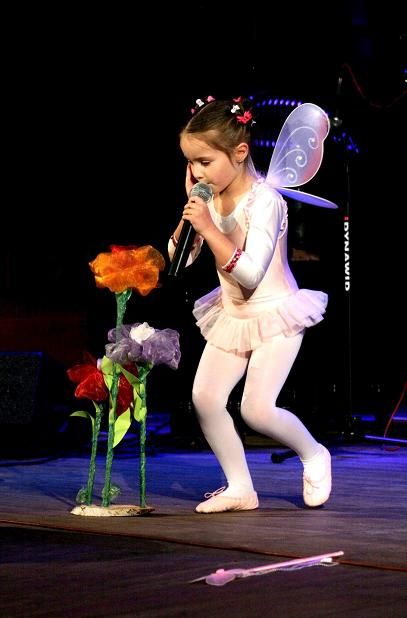 Dziewczynka przebrana za wróżkę śpiewa do mikrofonu, przed nią sztuczne kwiaty