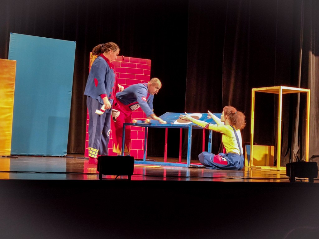 trójka aktorów rozmawia przy niebieskim stoliku na scenie