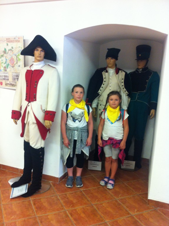 Dwie dziewczynki w żółtych chustach stojące przy trzech manekinach w strojach historycznych
